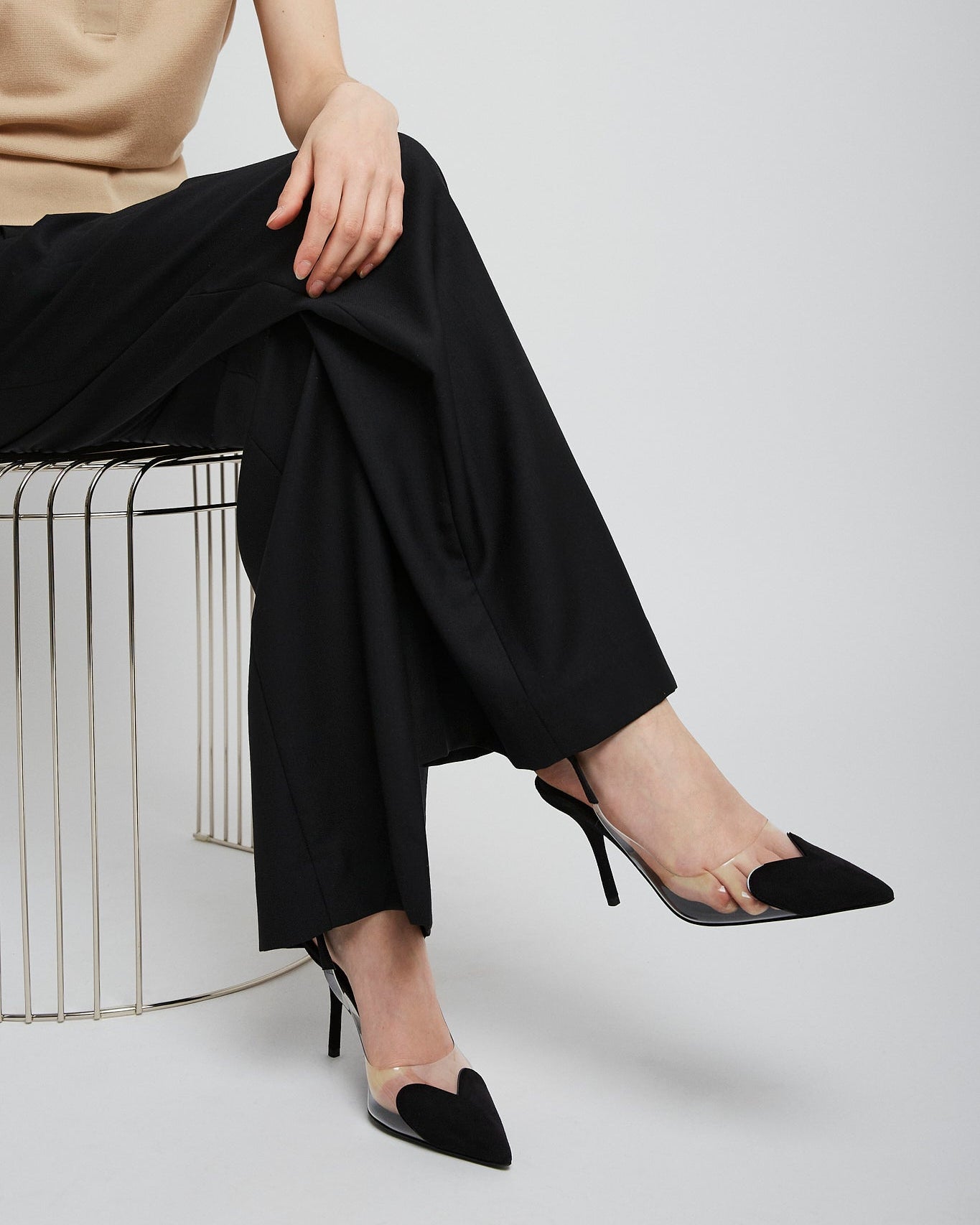 Person wearing black high-heeled shoes and black pants sitting on a white stool against a light gray background.