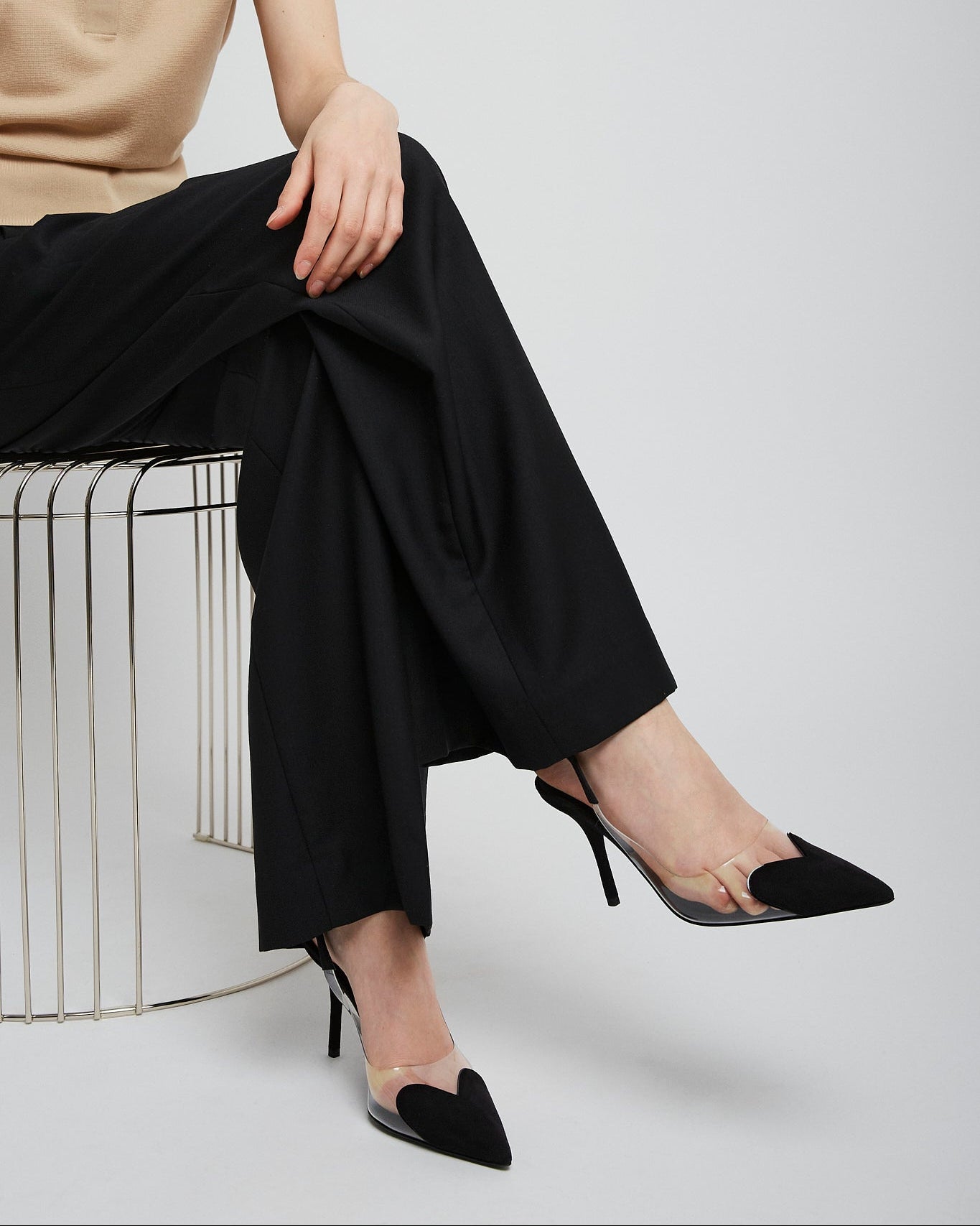 Person wearing black high-heeled shoes and black pants sitting on a white stool against a light gray background.