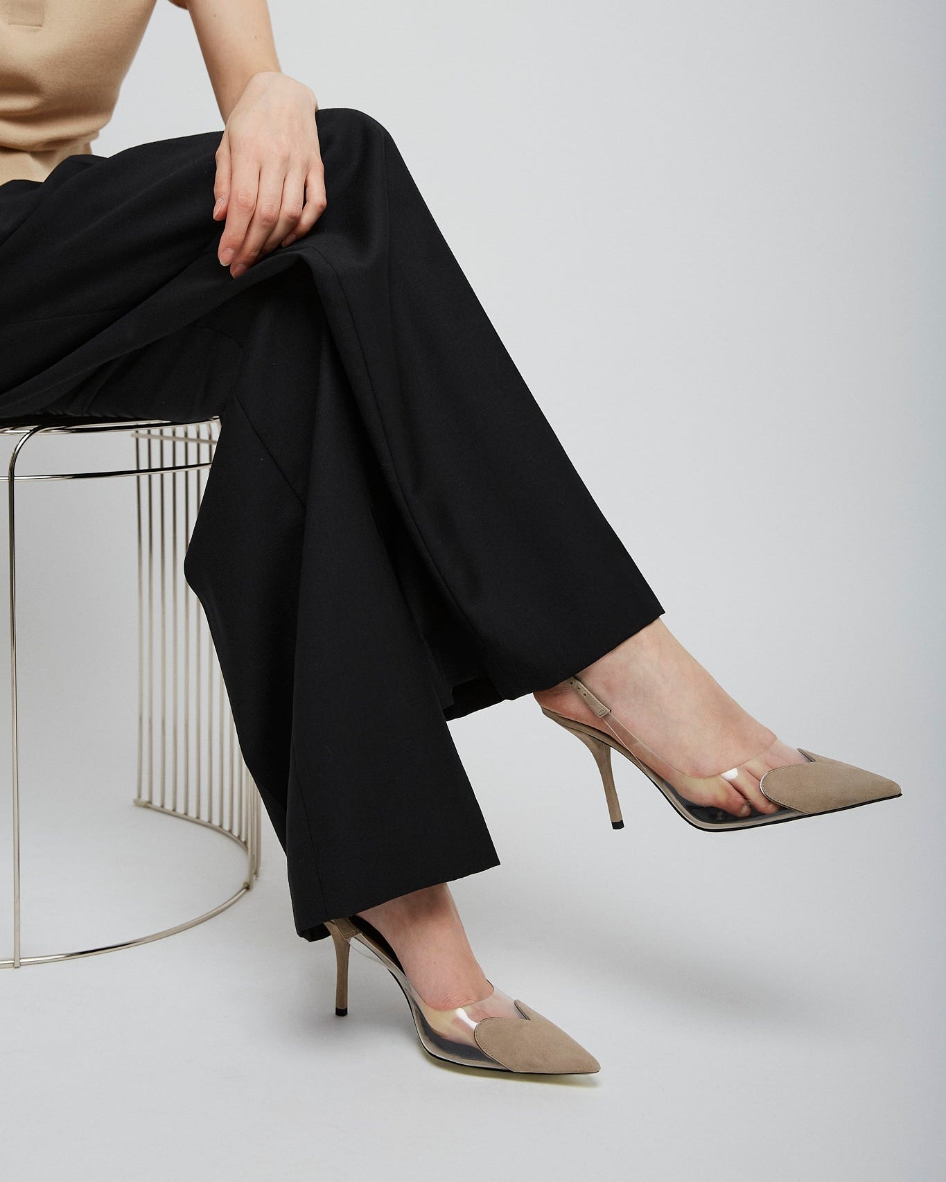 Person wearing black pants and beige high-heeled shoes sitting on a chair against a plain background