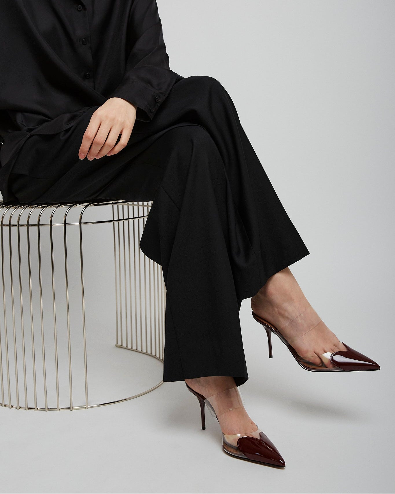 Person wearing a black outfit with high-heeled shoes sitting on a wire stool against a white background