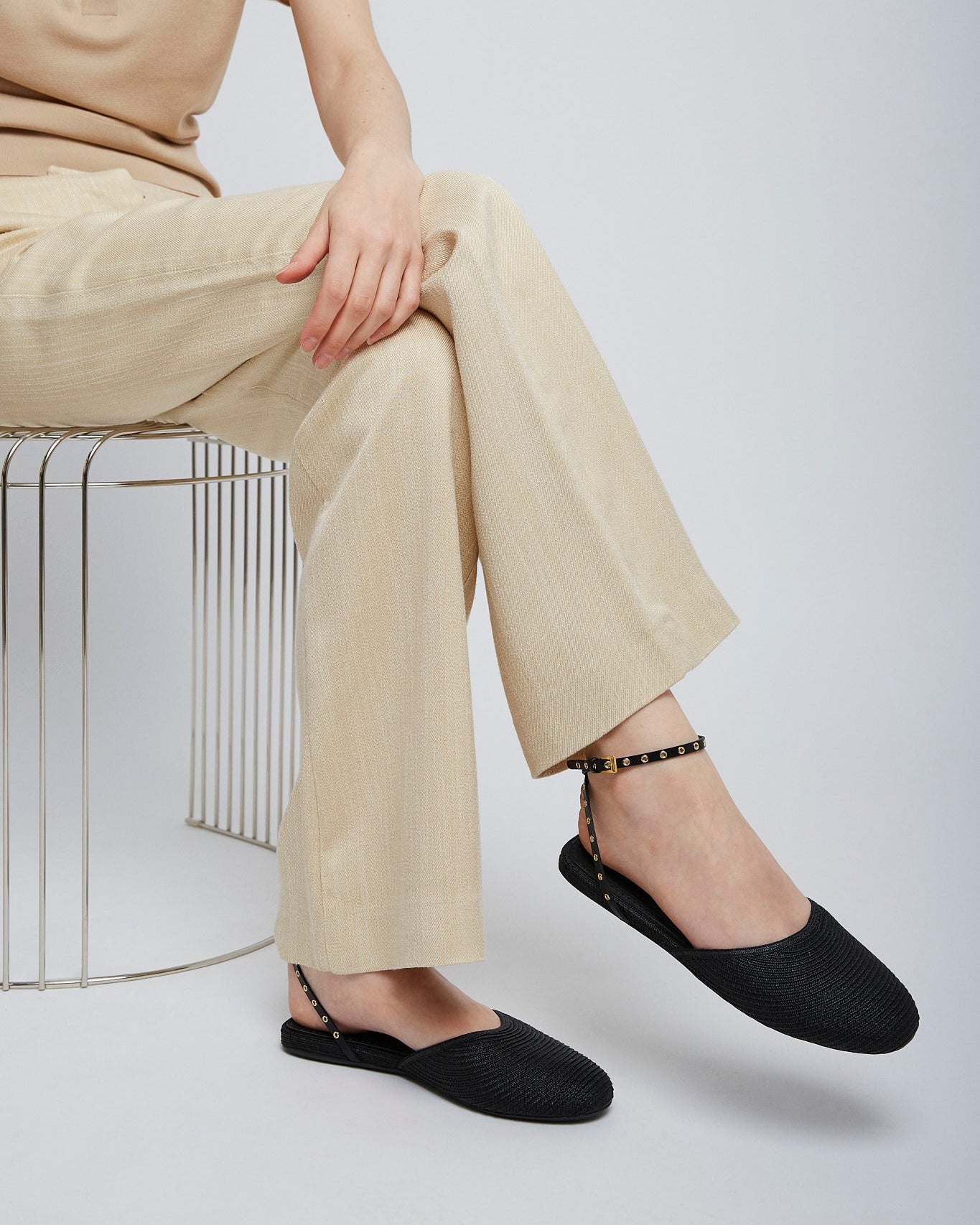 Person wearing beige pants and black shoes sitting on a wire stool against a white background