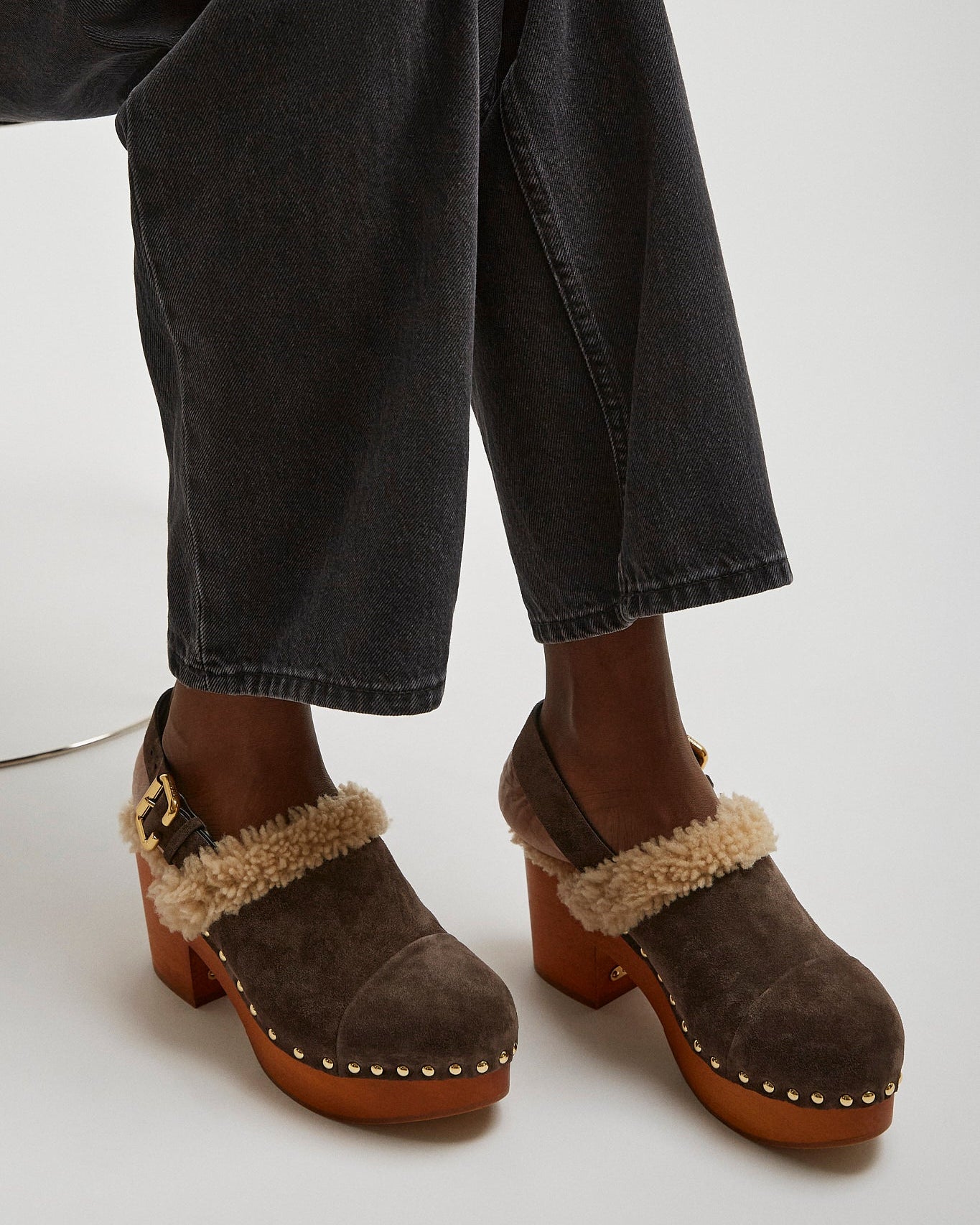 Brown suede clogs with fur trim worn with black jeans on a white background