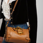 Brown leather handbag with gold accents worn by a person against a neutral background