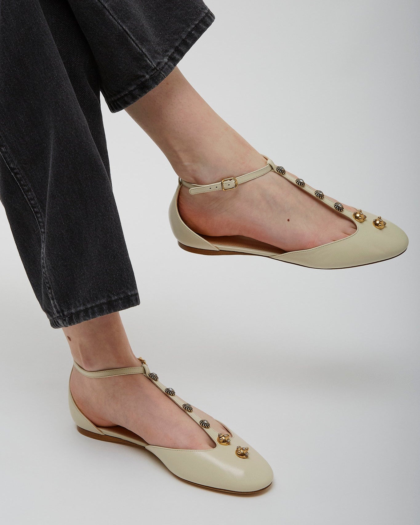 Beige flats with gold embellishments worn by a person in black pants on a white background