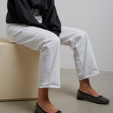 Person wearing a black shirt, white pants, and black studded flats sitting on a beige block.