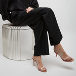 Person wearing a black outfit sitting on a wire stool against a white background