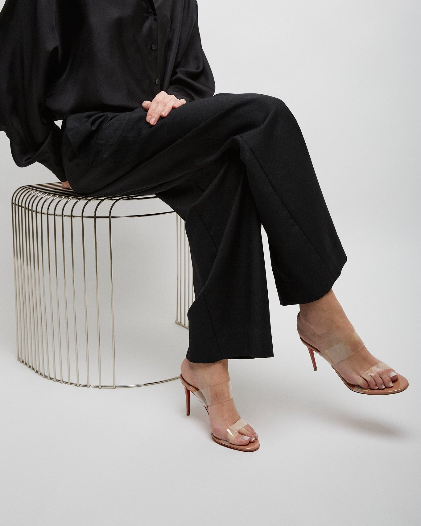 Person wearing a black outfit sitting on a wire stool against a white background
