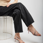 Person wearing black pants and transparent high-heeled sandals sitting on a wire stool against a white background.