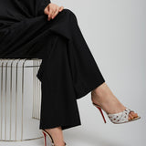 Person wearing black outfit and patterned high-heeled shoes sitting on a white stool.