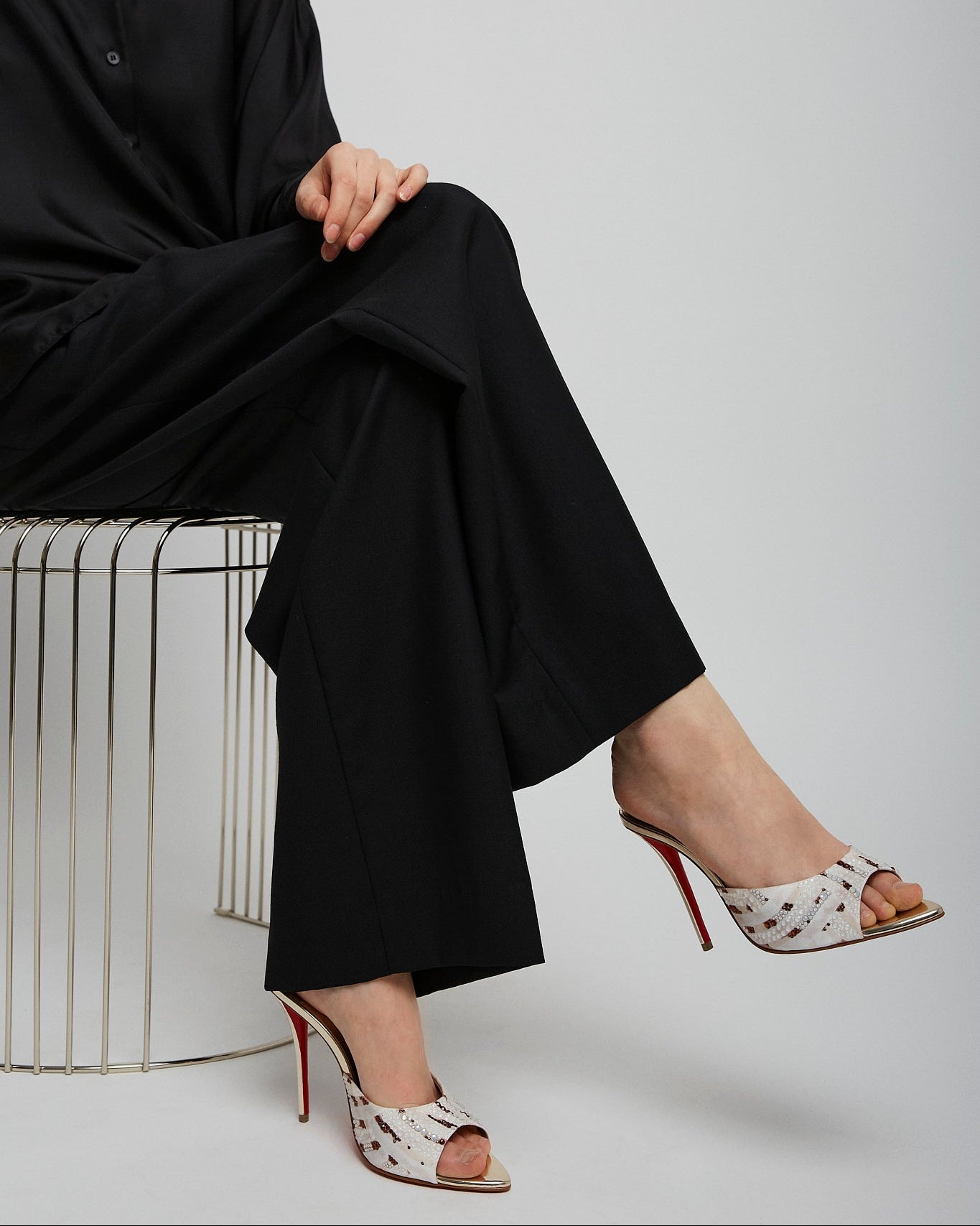 Person wearing black outfit and patterned high-heeled shoes sitting on a white stool.