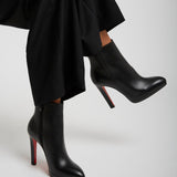 Black high-heeled boots with red soles worn by a person sitting on a light gray background