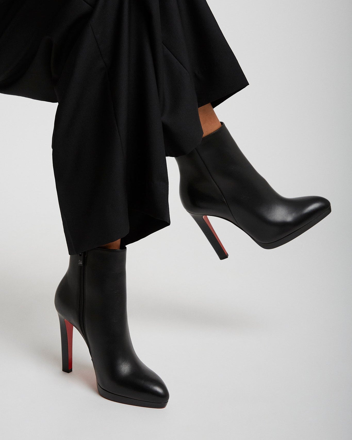Black high-heeled boots with red soles worn by a person sitting on a light gray background