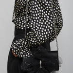 Person wearing a black and white polka dot blouse with a black handbag on a gray background