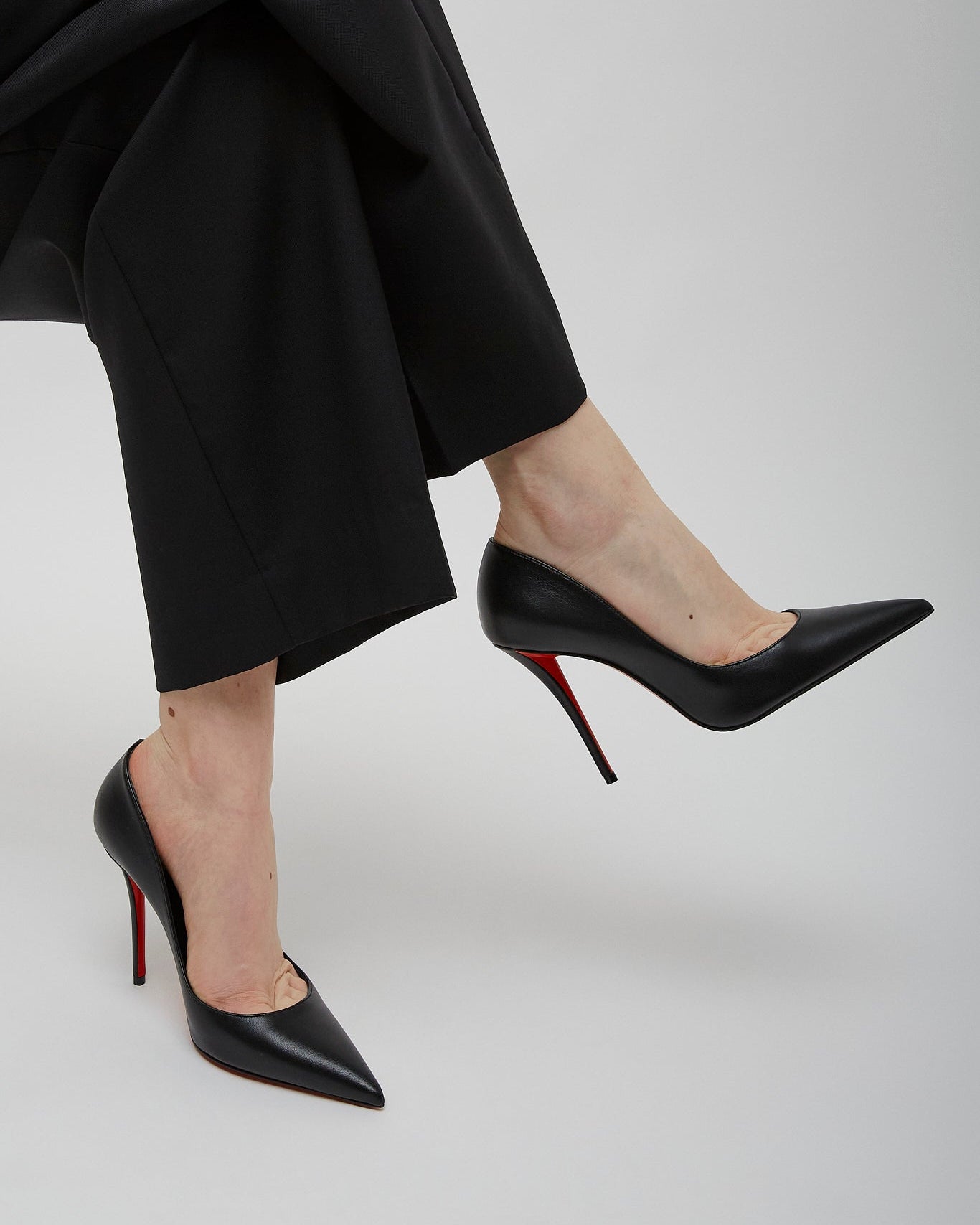 Person wearing black high-heeled shoes with red soles and black pants on a light gray background