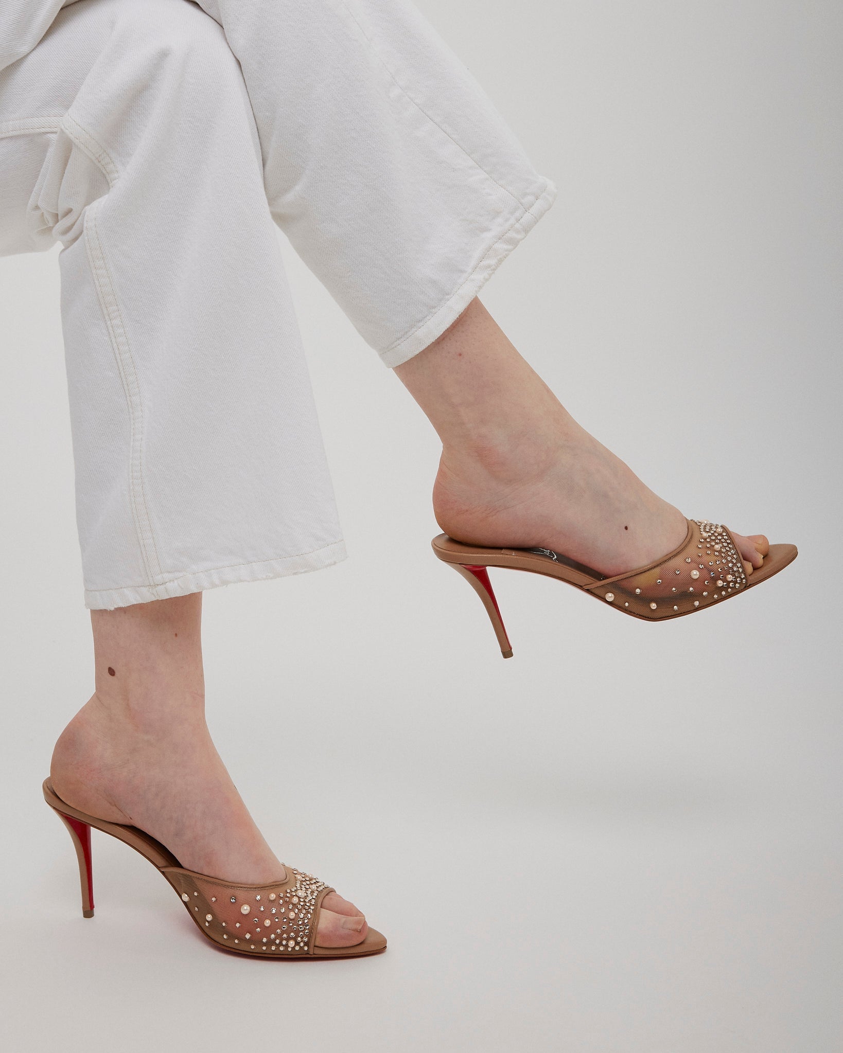 Gold high-heeled shoes with red soles worn by a person in white pants on a light gray background