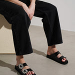 Person wearing black sandals with decorative straps sitting on a light-colored floor.