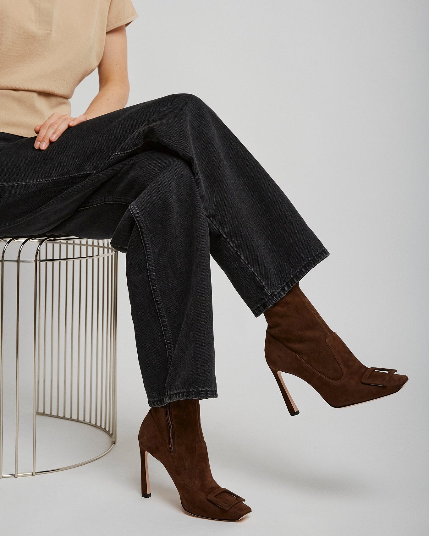 Person wearing a beige shirt, black jeans, and brown high-heeled shoes sitting on a wire stool.