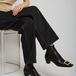 Person wearing black ankle boots with gold buckles, black jeans, and a beige shirt sitting on a wire stool.