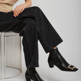 Person wearing black ankle boots with gold buckles, black jeans, and a beige shirt sitting on a wire stool.