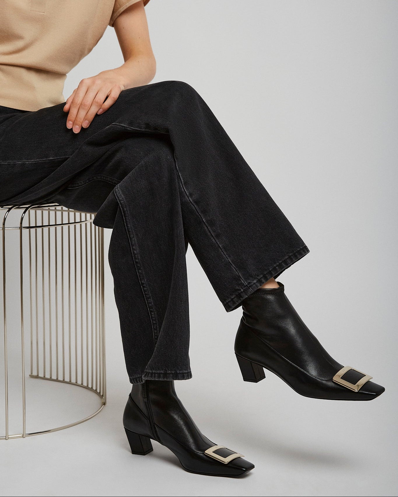 Person wearing black ankle boots with gold buckles, black jeans, and a beige shirt sitting on a wire stool.