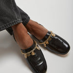 Black loafers with gold chain details worn with gray jeans on a light background