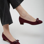 Burgundy shoes with a bow detail worn by a person in black pants on a white background