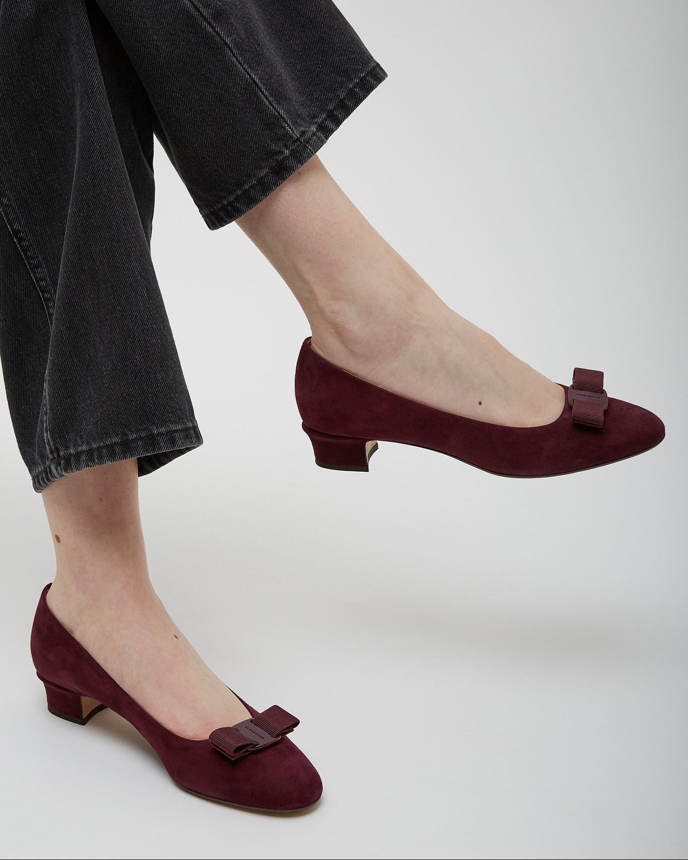 Burgundy shoes with a bow detail worn by a person in black pants on a white background