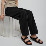 Person wearing a beige tank top and black pants sitting on a white stool against a plain background
