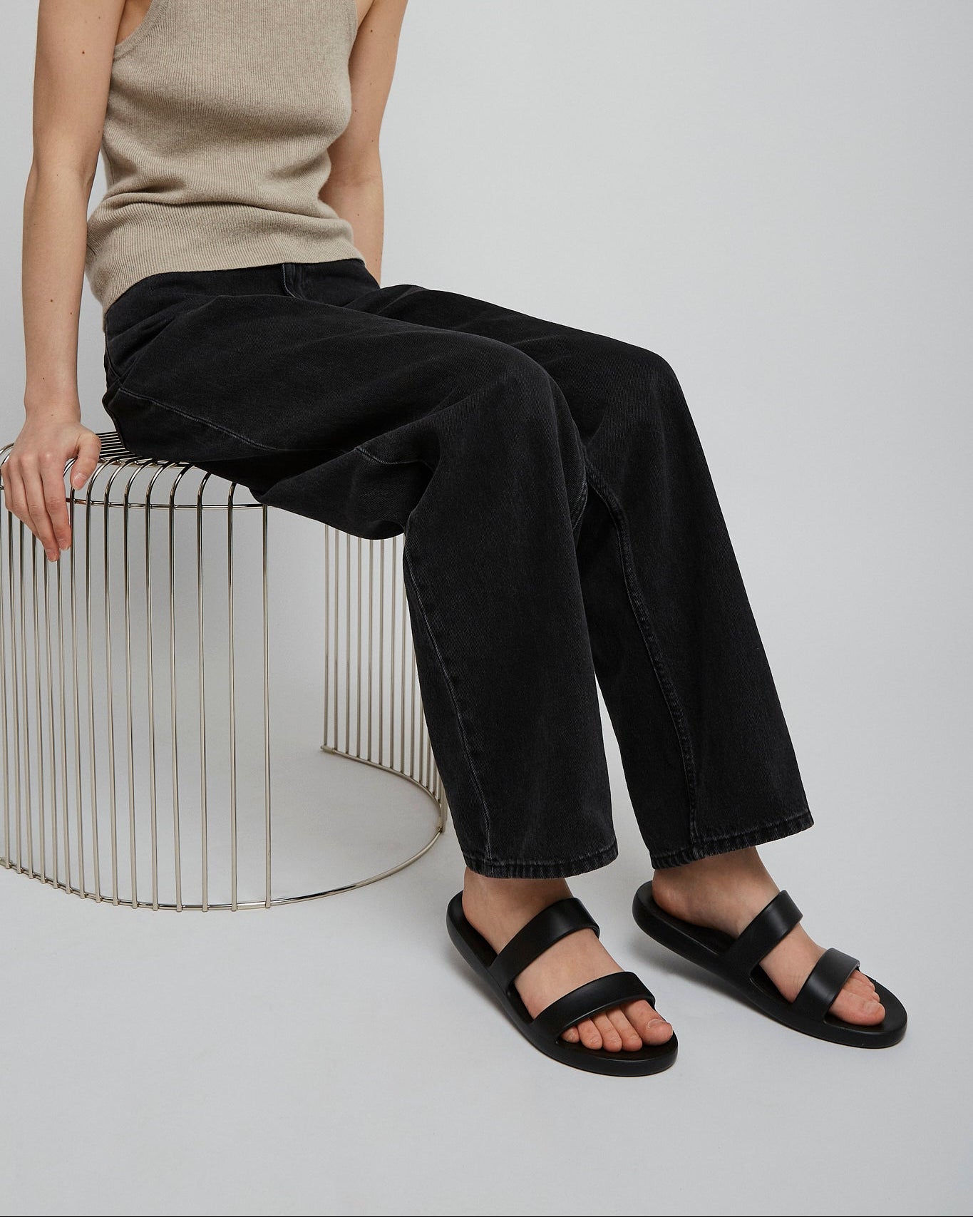 Person wearing a beige tank top and black pants sitting on a white stool against a plain background