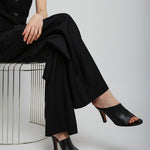 Person wearing a black outfit with high-heeled shoes sitting on a wire stool against a white background