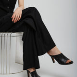 Person wearing a black outfit with high-heeled shoes sitting on a wire stool against a white background