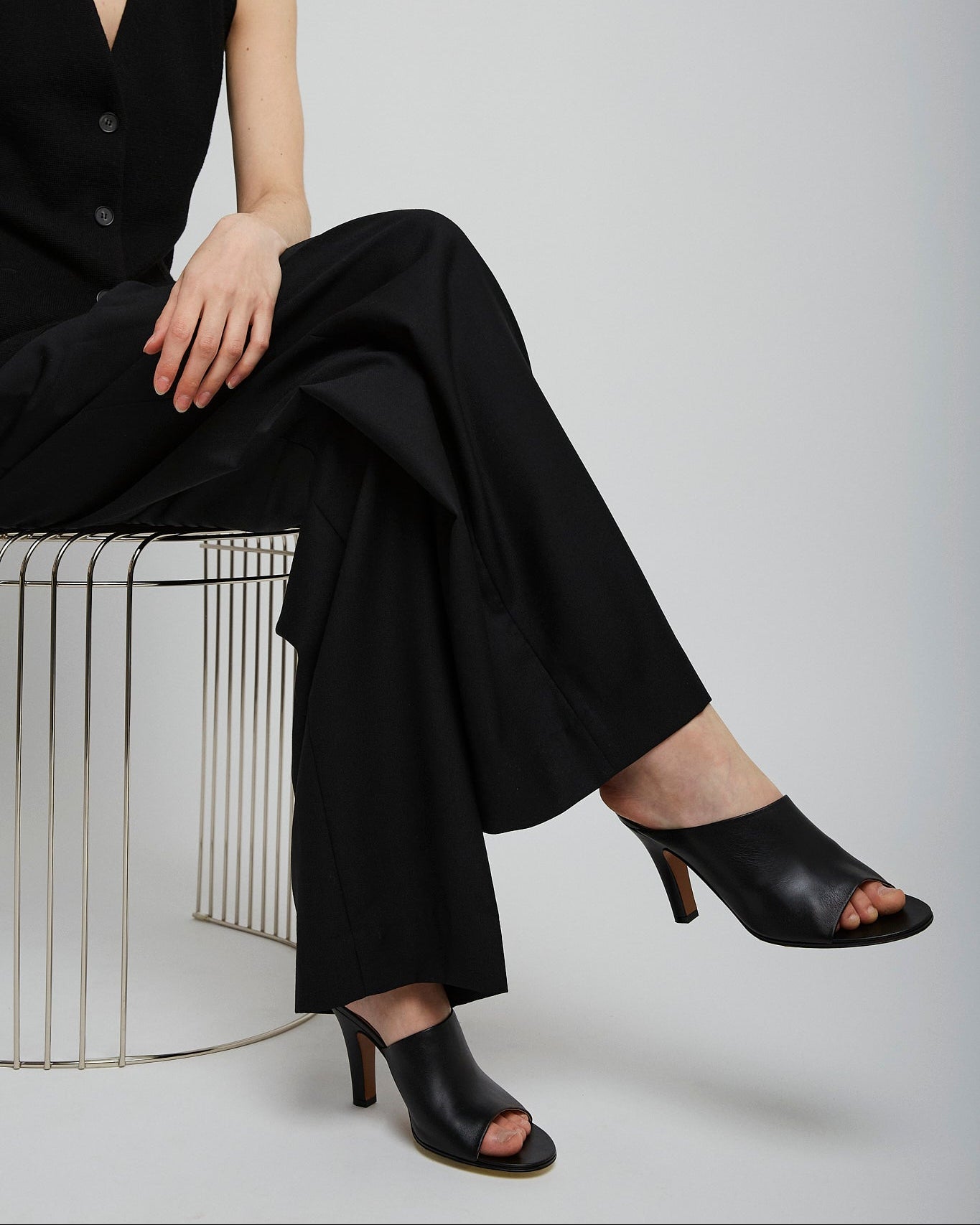 Person wearing a black outfit with high-heeled shoes sitting on a wire stool against a white background