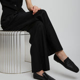 Person wearing a black sleeveless jumpsuit sitting on a metallic stool against a plain background