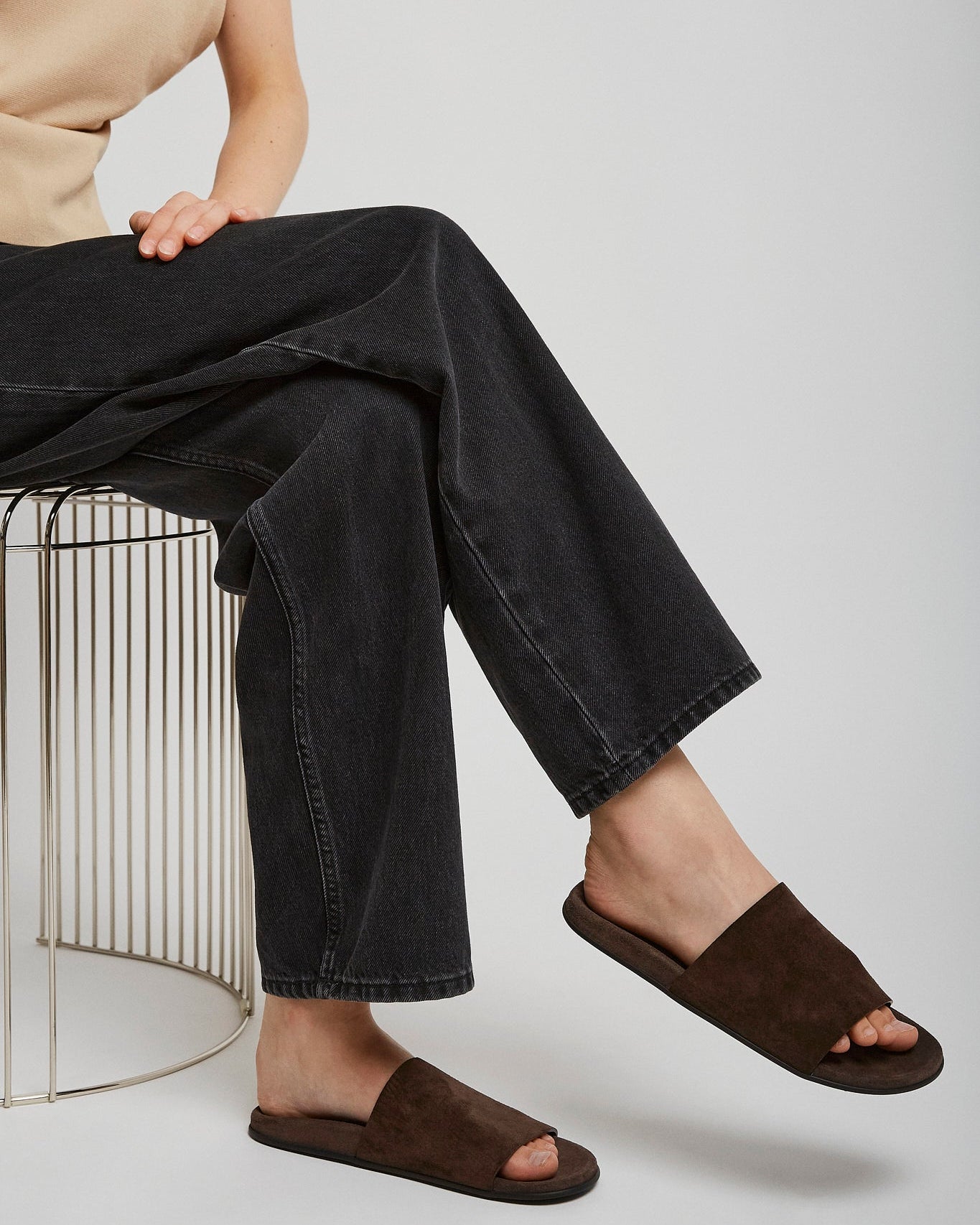 Person wearing brown sandals and black jeans sitting on a metal stool against a white background