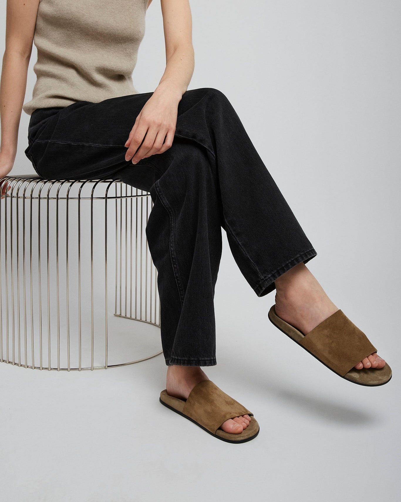 Person wearing a beige tank top, black pants, and brown sandals sitting on a white stool.