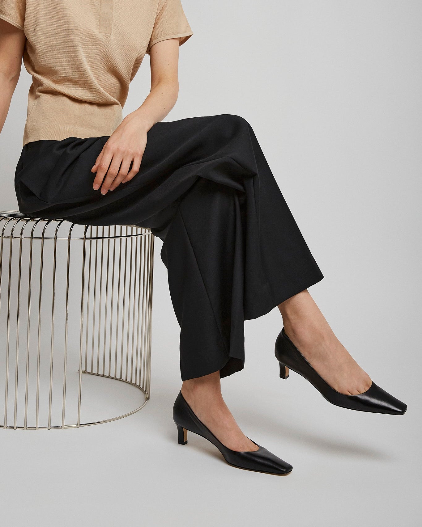 Person wearing a beige top and black pants sitting on a wire stool against a plain background