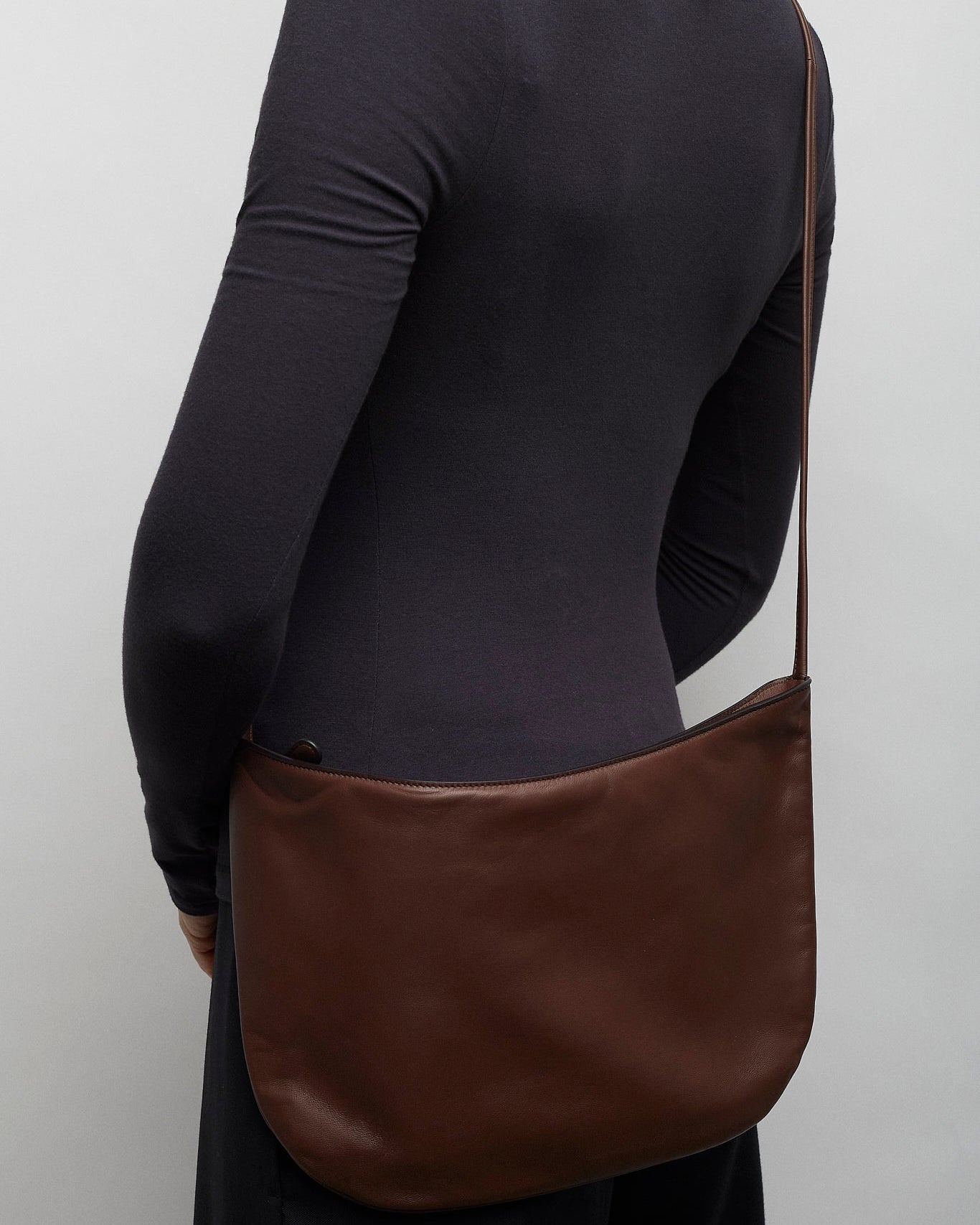 Person wearing a black long-sleeve shirt with a brown leather bag over one shoulder against a plain background.