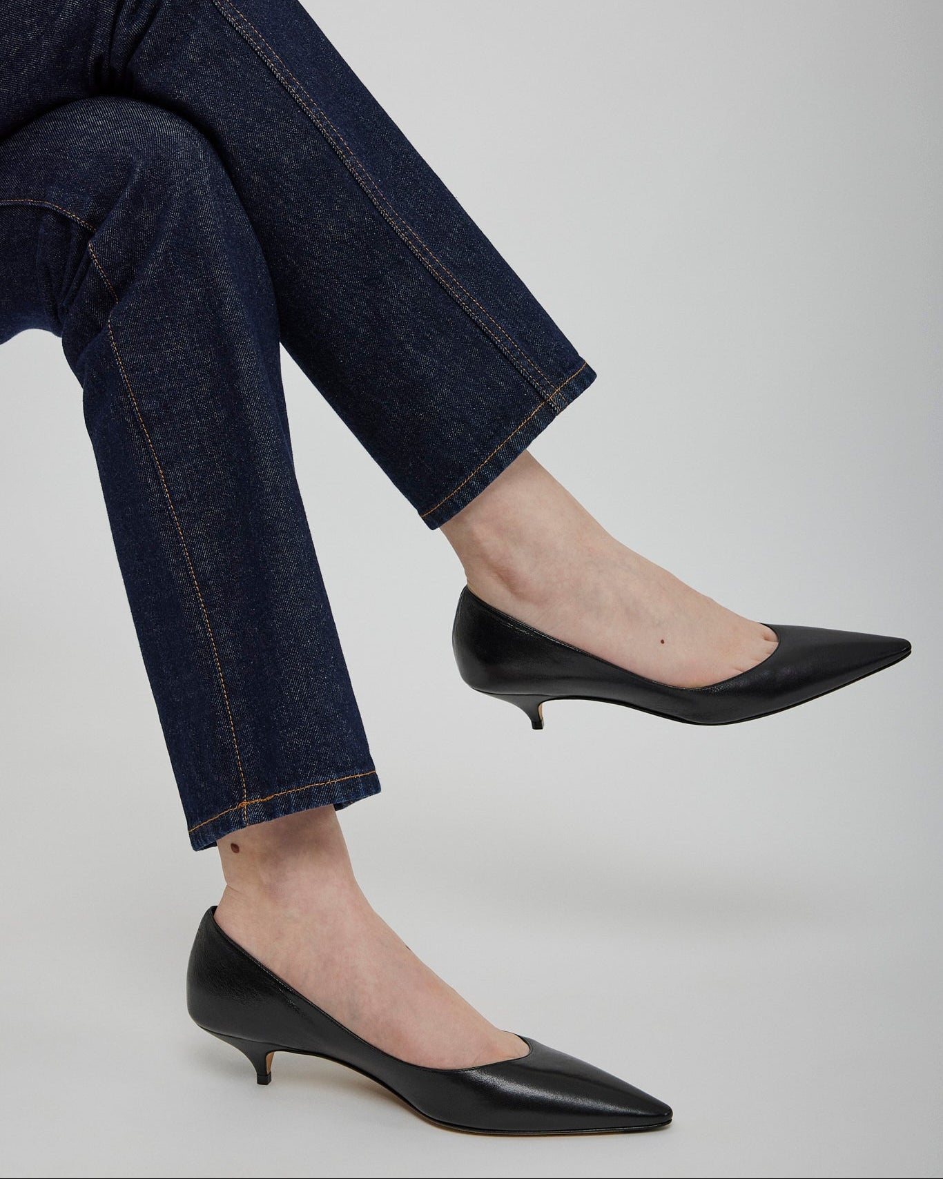 Person wearing dark blue jeans and black pointed-toe shoes on a light gray background