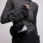 Person wearing a gray sweater with ruffled sleeves holding a black heart-shaped bag.