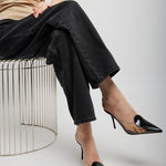 Person wearing black pants and black high-heeled shoes sitting on a metal stool against a plain background