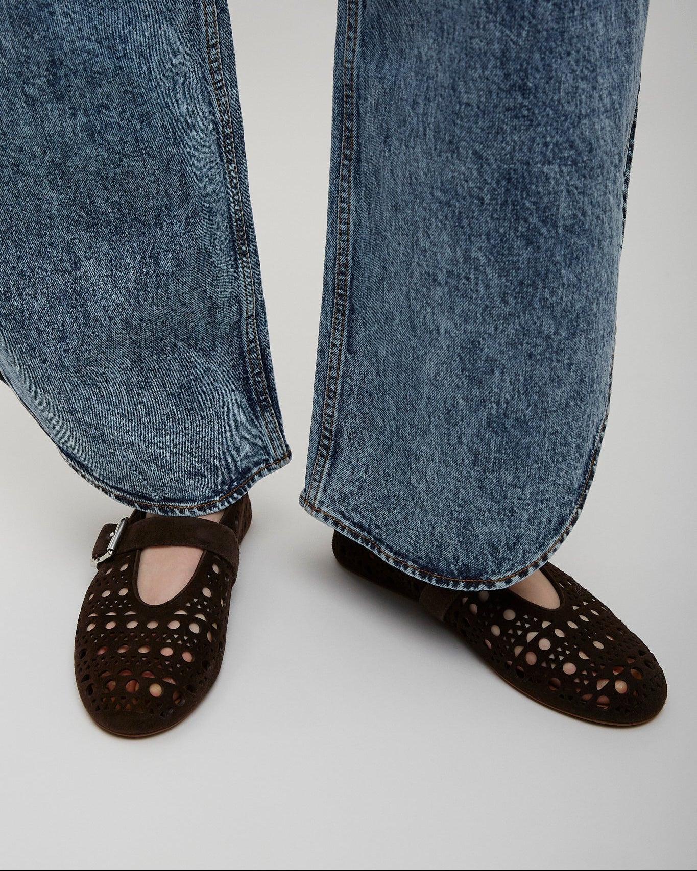 Person wearing blue jeans and brown perforated shoes on a light gray background