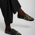 Black loafers with gold buckles worn by a person in black pants on a light gray background