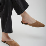 Person wearing brown suede slippers and black jeans on a light gray background