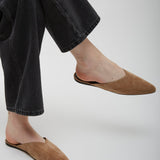 Person wearing brown suede slippers and black jeans on a light gray background