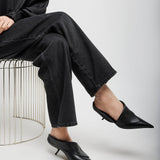 Person wearing black outfit with high-waisted pants and pointed-toe shoes sitting on a wire stool.