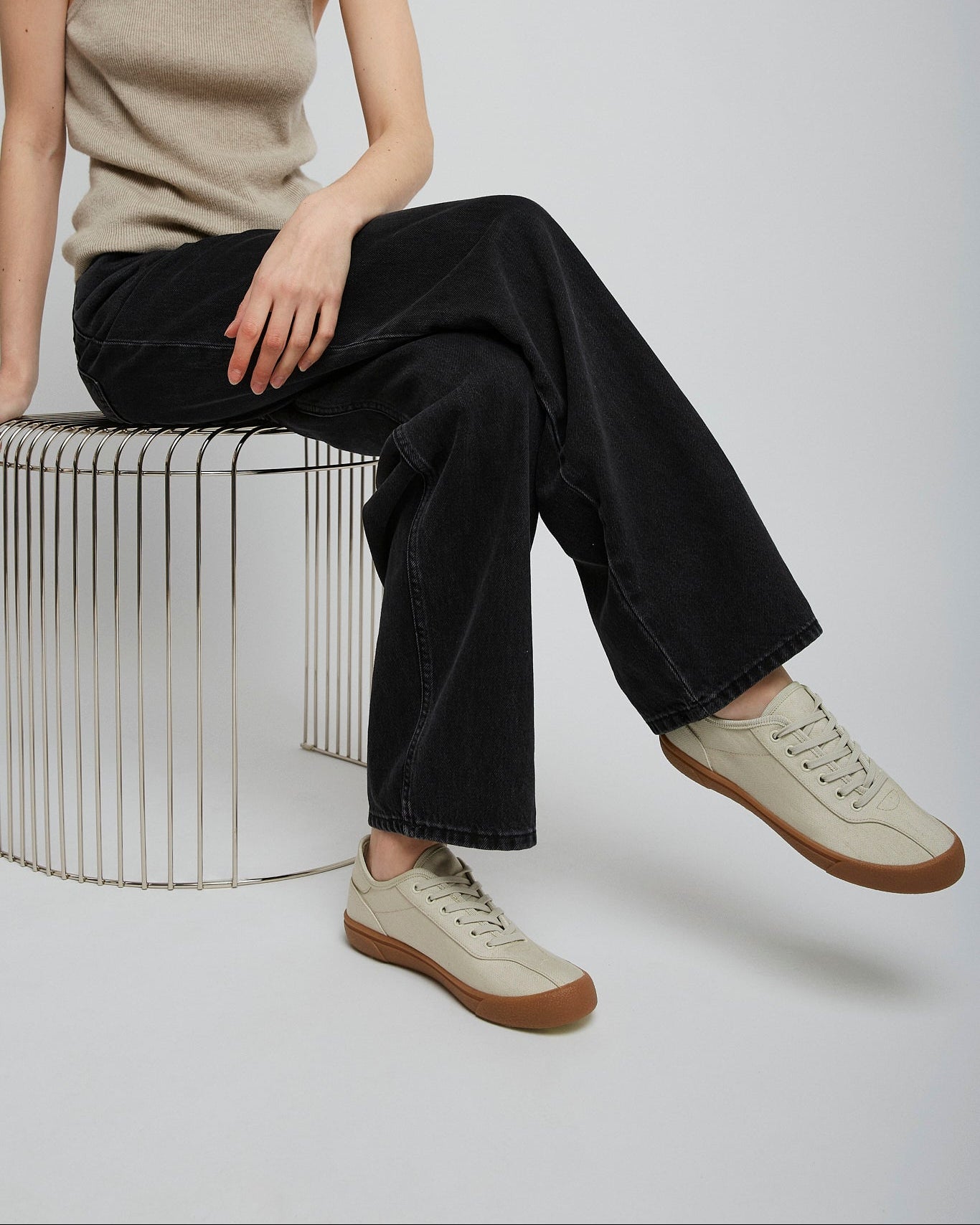 Person wearing a beige tank top, black pants, and beige sneakers sitting on a metal stool.